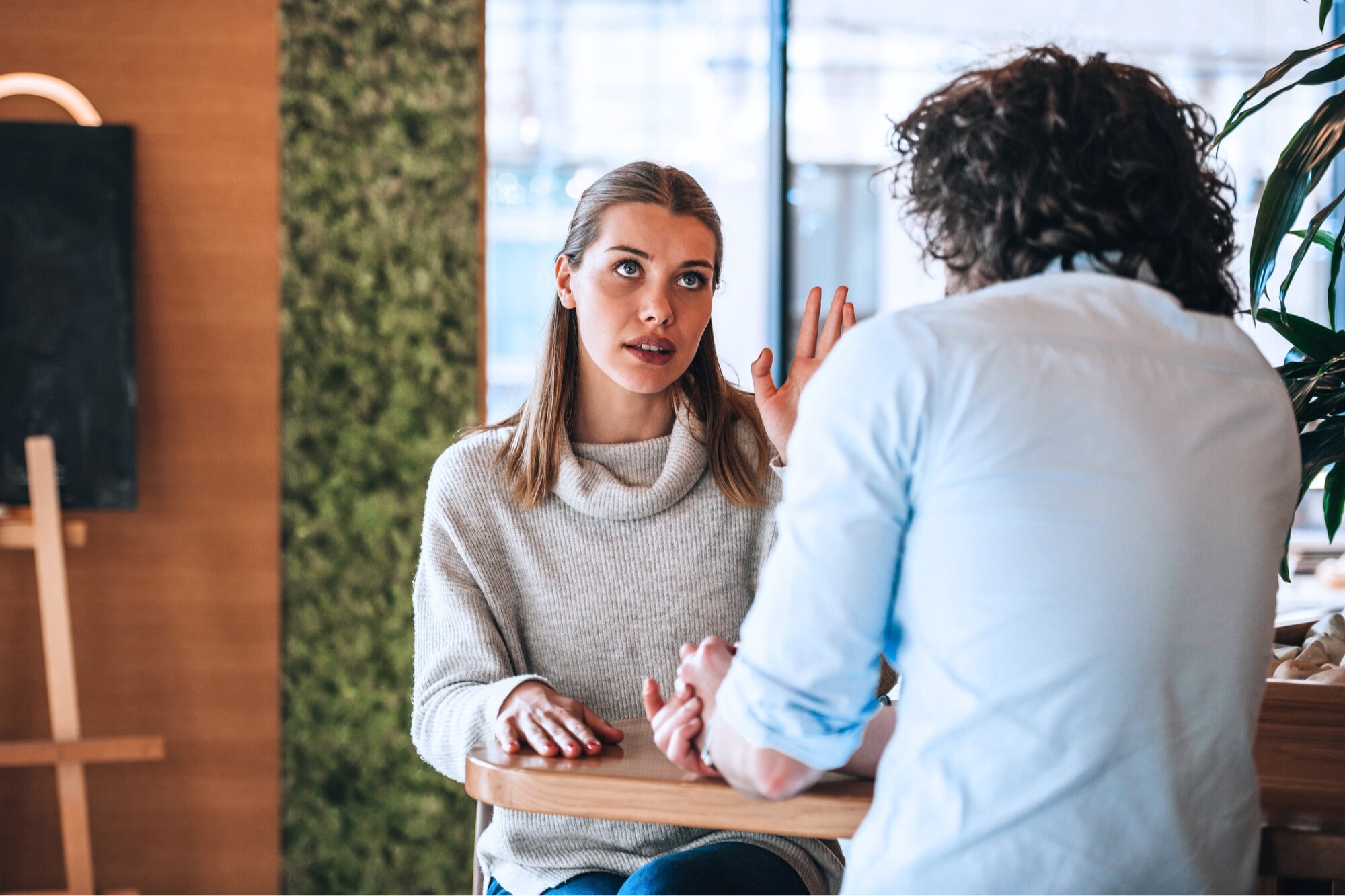 Two people are sitting at a wooden table in a conversation. The woman, wearing a light sweater, appears to be speaking with an expressive gesture, while the man, with curly hair, listens attentively. The setting is a cozy indoor space.