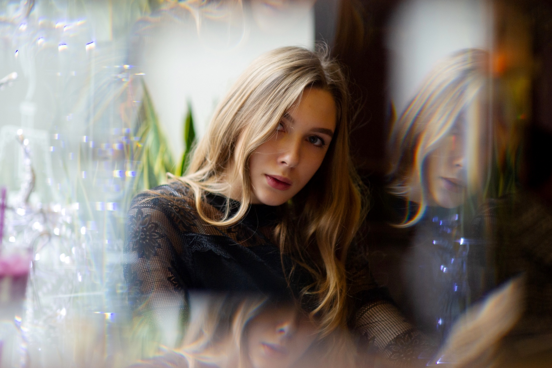 A woman with long blonde hair, wearing a black lace top, is sitting in a reflective setting. Soft lighting creates a dreamy, blurred effect with her reflection visible in the background.