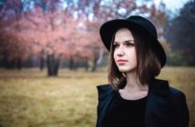 A woman wearing a black hat and coat stands in a field with autumn trees in the background. The scene is serene, with soft pink and brown hues from the leaves, creating a calm, contemplative atmosphere.