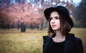 A woman wearing a black hat and coat stands in a field with autumn trees in the background. The scene is serene, with soft pink and brown hues from the leaves, creating a calm, contemplative atmosphere.