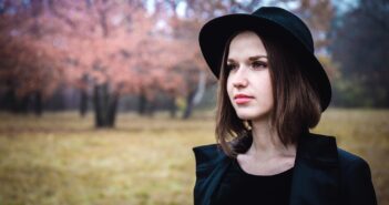 A woman wearing a black hat and coat stands in a field with autumn trees in the background. The scene is serene, with soft pink and brown hues from the leaves, creating a calm, contemplative atmosphere.