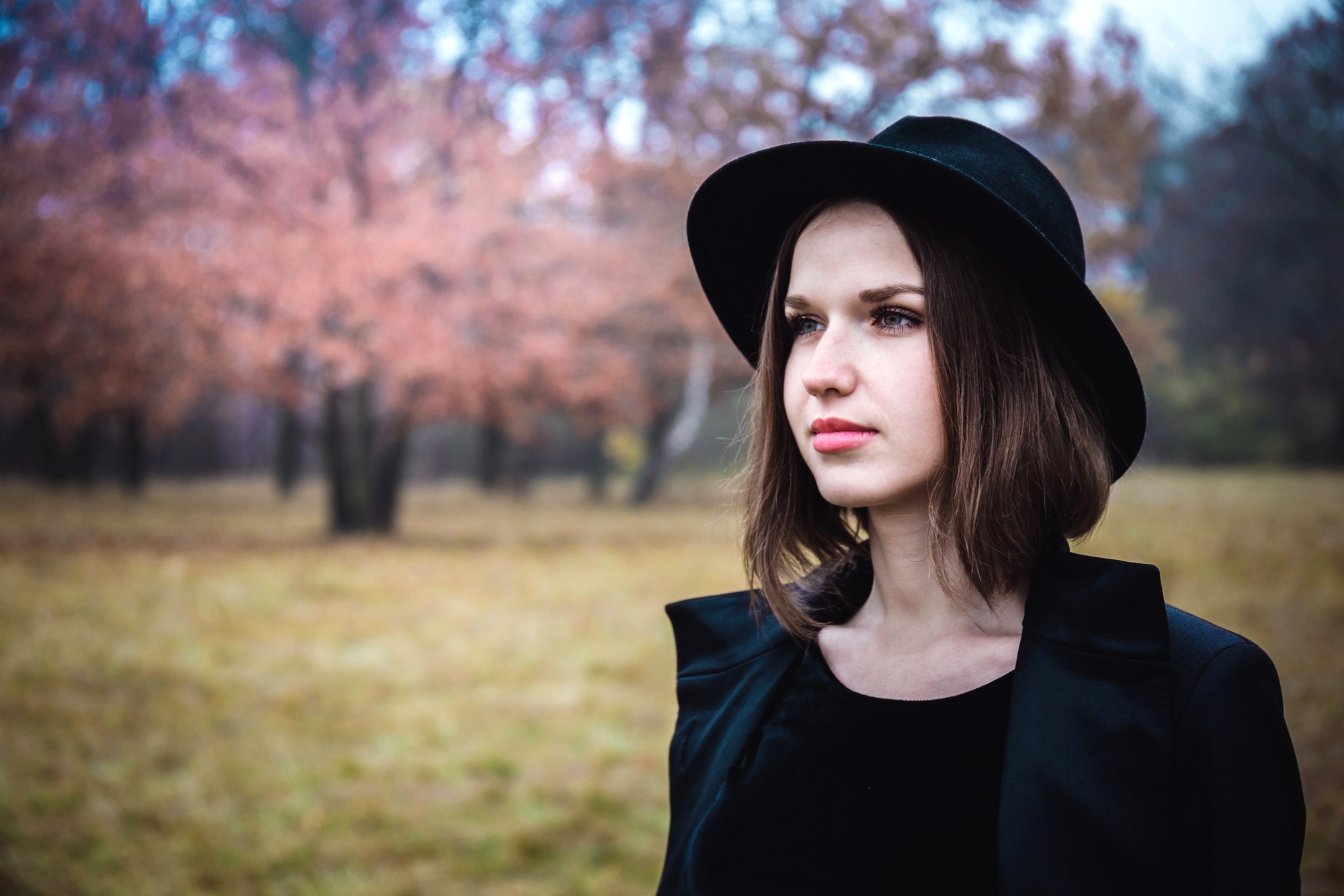 A woman wearing a black hat and coat stands in a field with autumn trees in the background. The scene is serene, with soft pink and brown hues from the leaves, creating a calm, contemplative atmosphere.