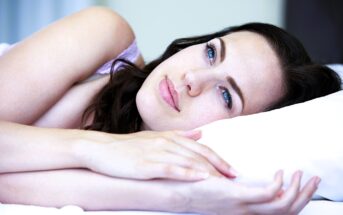 A woman with dark hair is lying on her side, resting her head on a white pillow. She has a calm expression, looking up with blue eyes. The background is softly blurred, highlighting her face.