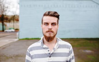 A person with a beard and blue eyes stands outside in front of a light blue wall, wearing a gray and white striped shirt.