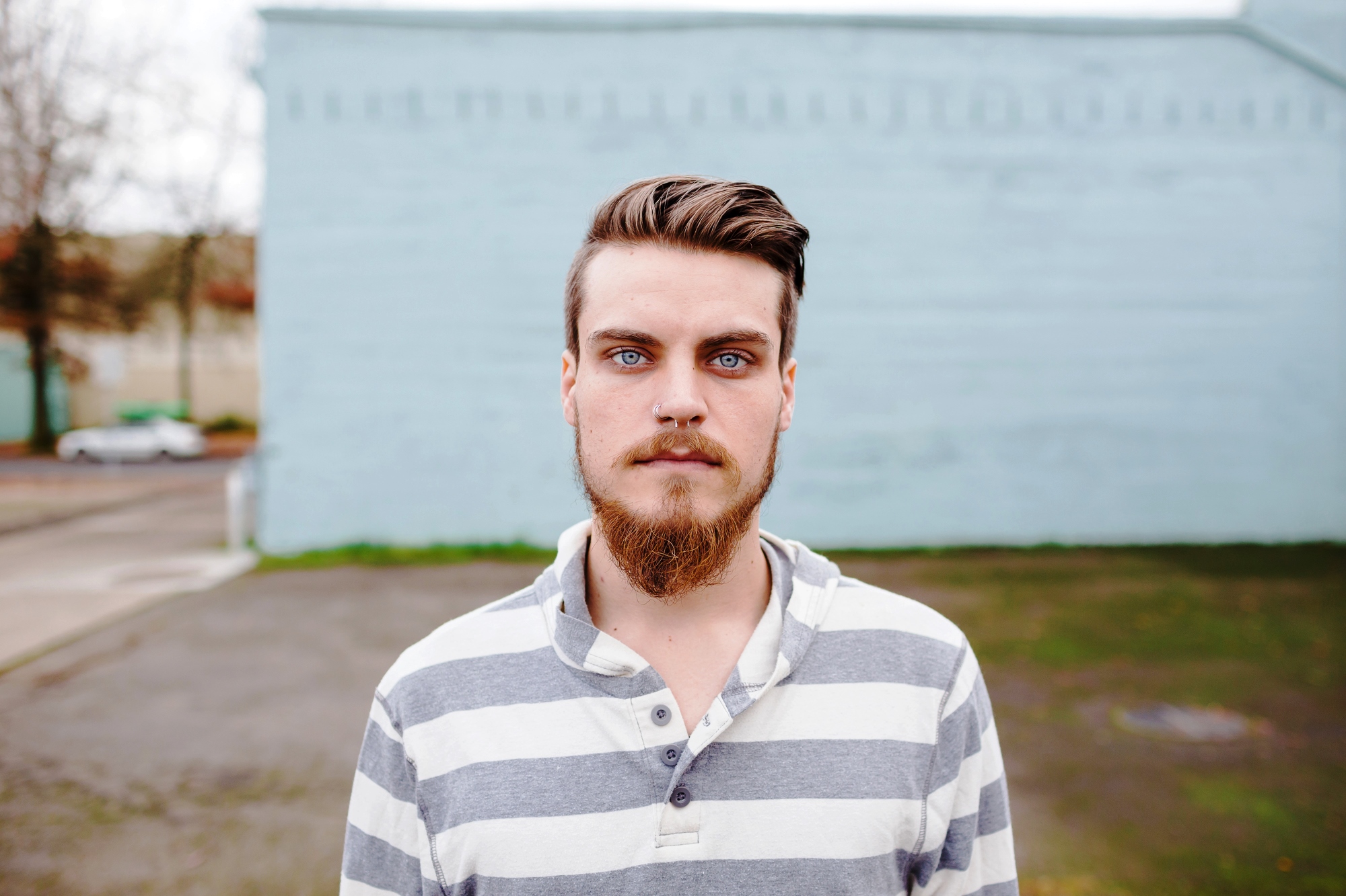 A person with a beard and blue eyes stands outside in front of a light blue wall, wearing a gray and white striped shirt.