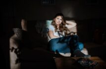 A woman sitting on a sofa in a dimly lit room, with sunlight creating patterns on her and the furniture. She wears a white shirt and jeans, resting her head on her hand. A book is on the wooden table in the foreground.