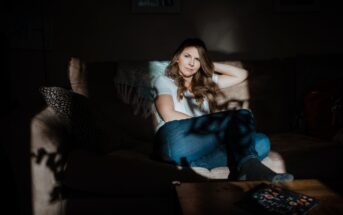 A woman sitting on a sofa in a dimly lit room, with sunlight creating patterns on her and the furniture. She wears a white shirt and jeans, resting her head on her hand. A book is on the wooden table in the foreground.