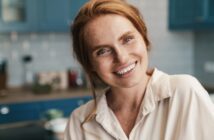 A person with long red hair smiles warmly and is wearing a light-colored shirt in a kitchen setting with blue cabinets and a blurred background.