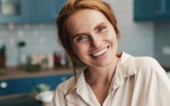 A person with long red hair smiles warmly and is wearing a light-colored shirt in a kitchen setting with blue cabinets and a blurred background.