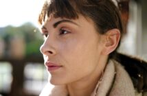 A person with short brown hair gazes thoughtfully into the distance. They are wearing a light-colored scarf and are illuminated by soft, natural light, with a blurred outdoor background.