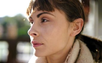 A person with short brown hair gazes thoughtfully into the distance. They are wearing a light-colored scarf and are illuminated by soft, natural light, with a blurred outdoor background.