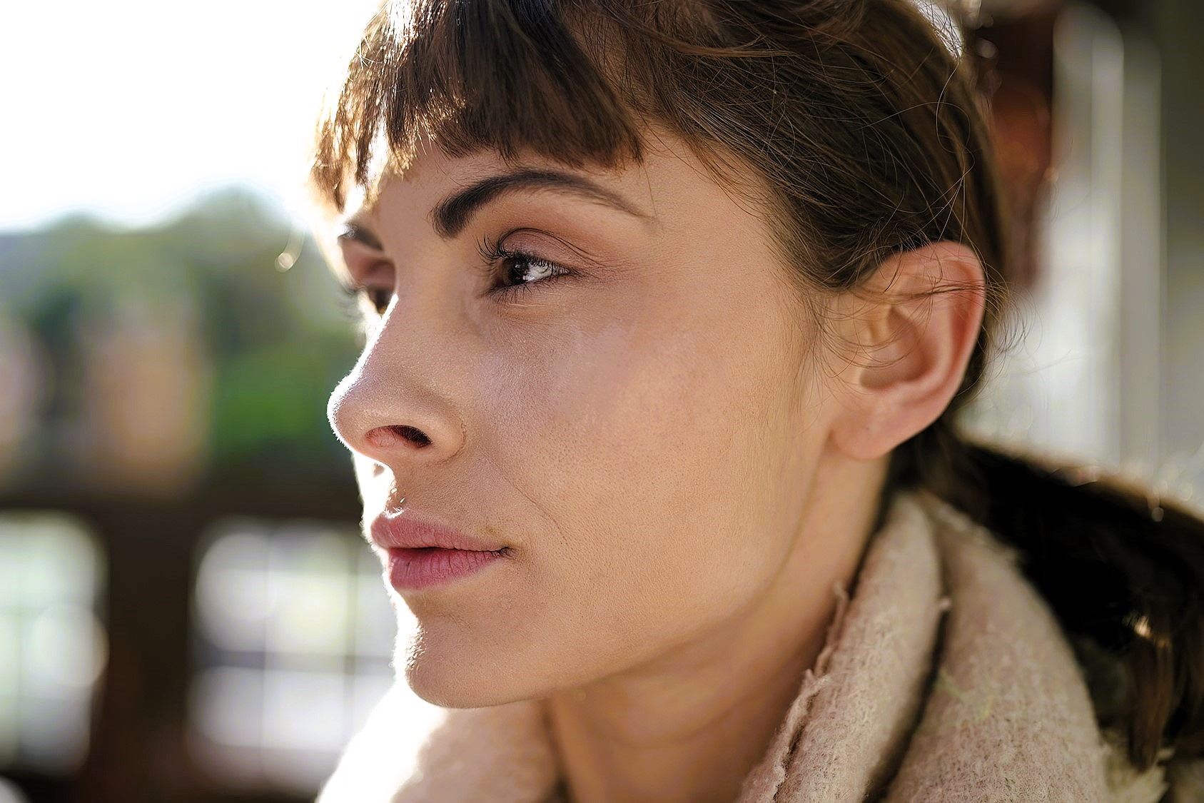 A person with short brown hair gazes thoughtfully into the distance. They are wearing a light-colored scarf and are illuminated by soft, natural light, with a blurred outdoor background.