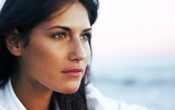 A woman with long dark hair gazes thoughtfully into the distance. She has a serene expression and is outdoors with a soft-focus background that suggests a calm setting.