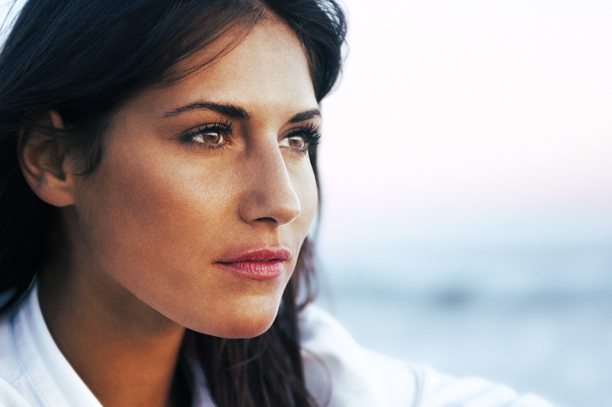 A woman with long dark hair gazes thoughtfully into the distance. She has a serene expression and is outdoors with a soft-focus background that suggests a calm setting.