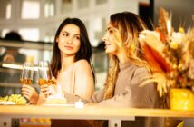 Two women sitting at a table, each holding a wine glass. One woman is speaking while the other listens. There are plates with appetizers and a vase of dried flowers on the table. The setting appears cozy and warmly lit.