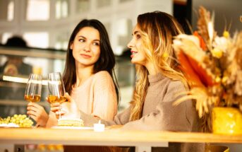 Two women sitting at a table, each holding a wine glass. One woman is speaking while the other listens. There are plates with appetizers and a vase of dried flowers on the table. The setting appears cozy and warmly lit.