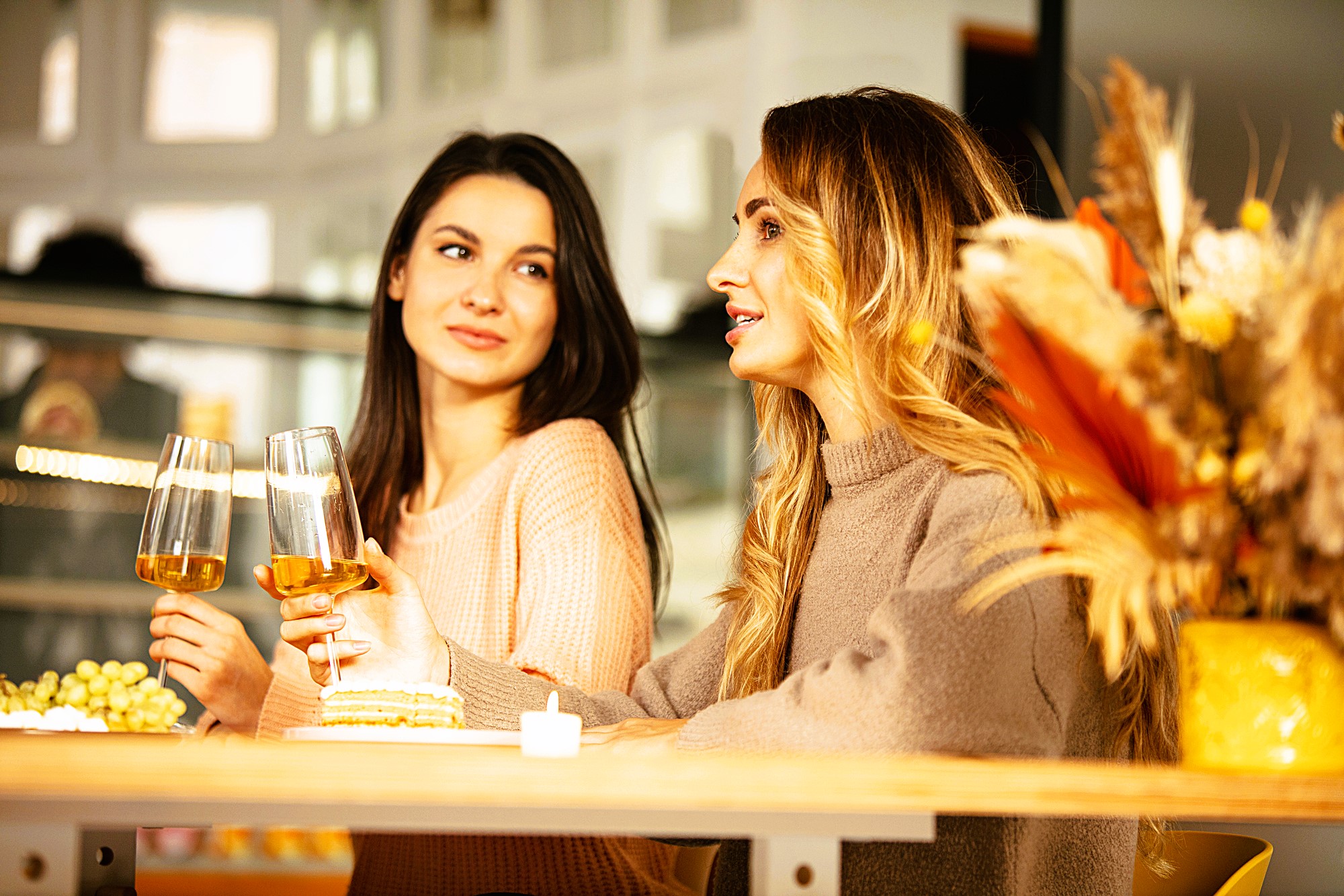 Two women sitting at a table, each holding a wine glass. One woman is speaking while the other listens. There are plates with appetizers and a vase of dried flowers on the table. The setting appears cozy and warmly lit.