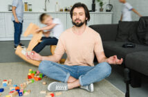 A man sits cross-legged on the floor, meditating amidst colorful toy blocks. Two children play energetically in the background, one on a rocking horse. The living room is light and modern, with a dark sofa and kitchen space visible.