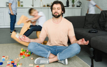 A man sits cross-legged on the floor, meditating amidst colorful toy blocks. Two children play energetically in the background, one on a rocking horse. The living room is light and modern, with a dark sofa and kitchen space visible.