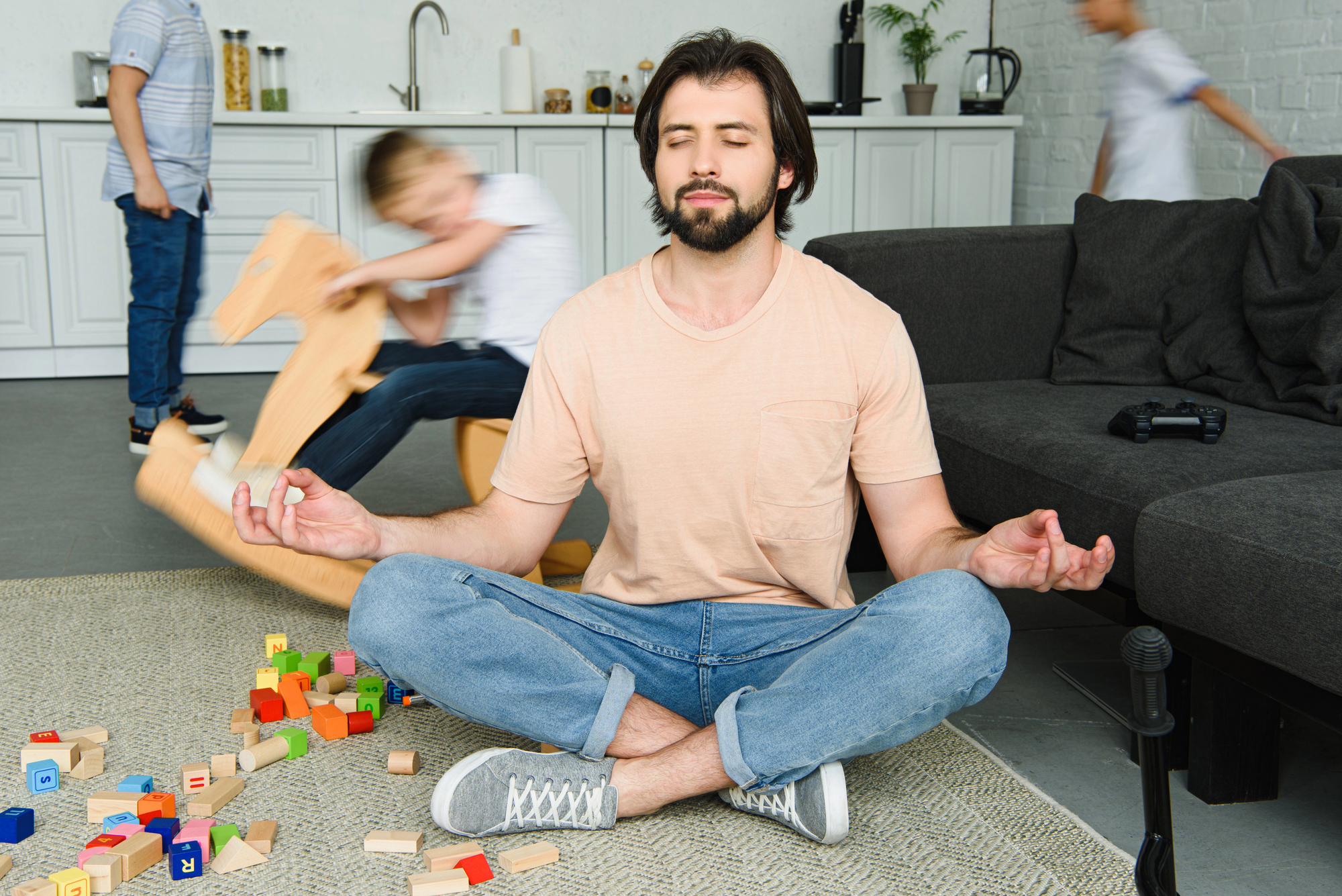 A man sits cross-legged on the floor, meditating amidst colorful toy blocks. Two children play energetically in the background, one on a rocking horse. The living room is light and modern, with a dark sofa and kitchen space visible.