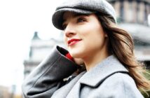 Woman in a gray coat and hat stands outdoors, looking upward with a slight smile. The background is a blurred cityscape, suggesting an urban environment.