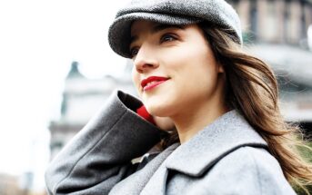 Woman in a gray coat and hat stands outdoors, looking upward with a slight smile. The background is a blurred cityscape, suggesting an urban environment.