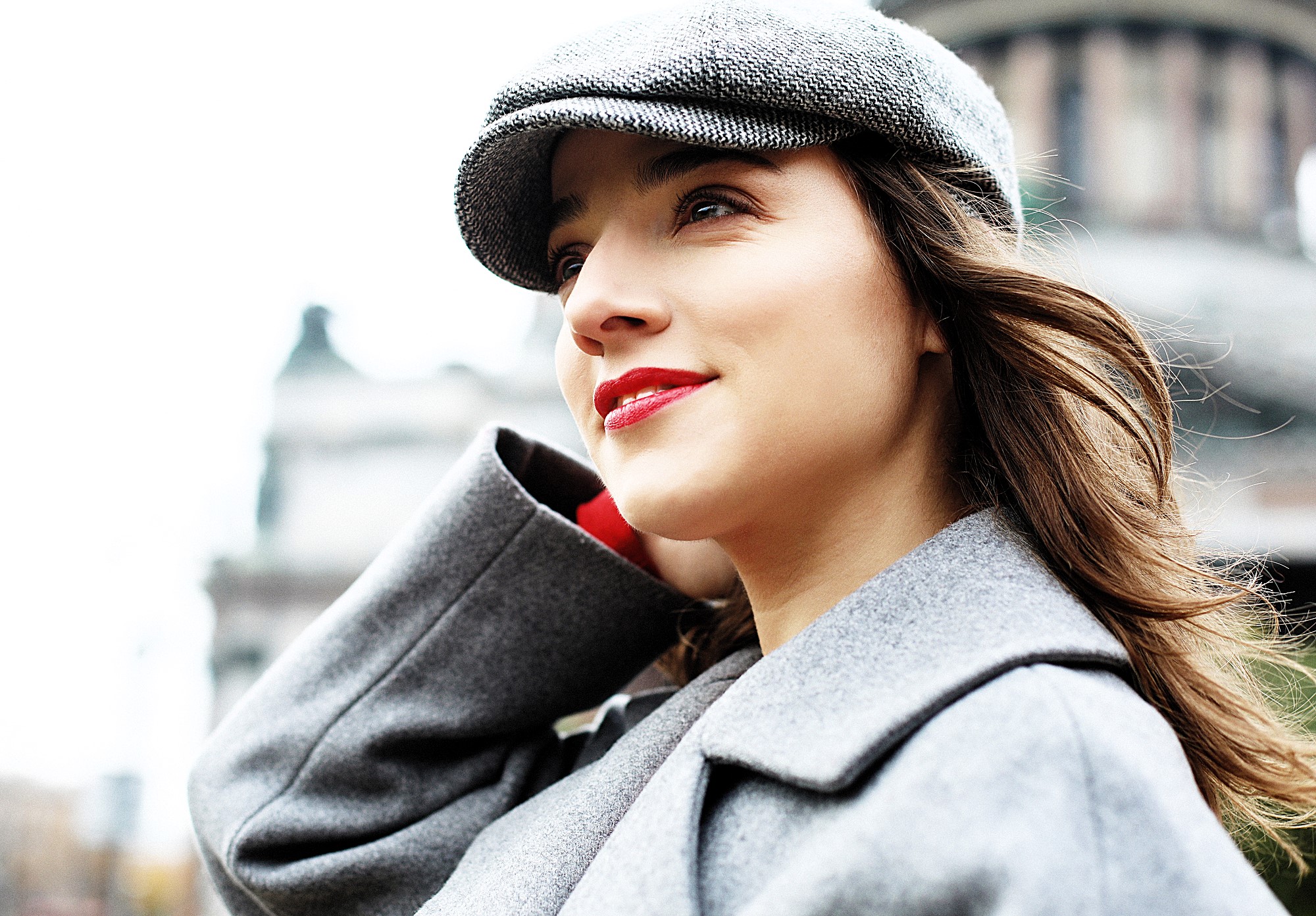 Woman in a gray coat and hat stands outdoors, looking upward with a slight smile. The background is a blurred cityscape, suggesting an urban environment.