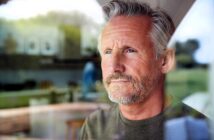 A contemplative man with gray hair and a beard looks out of a window. He is indoors, and the background shows a blurred view of a living space with natural light filtering through.