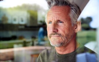 A contemplative man with gray hair and a beard looks out of a window. He is indoors, and the background shows a blurred view of a living space with natural light filtering through.