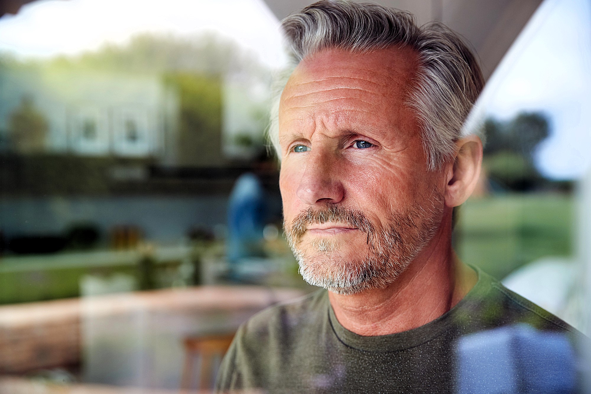 A contemplative man with gray hair and a beard looks out of a window. He is indoors, and the background shows a blurred view of a living space with natural light filtering through.