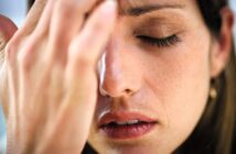 A close-up of a person with closed eyes, gently touching their forehead with one hand. They appear to be experiencing discomfort or deep thought. Their expression is calm, with a focus on their fingers and facial features.