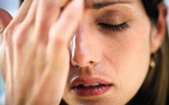 A close-up of a person with closed eyes, gently touching their forehead with one hand. They appear to be experiencing discomfort or deep thought. Their expression is calm, with a focus on their fingers and facial features.