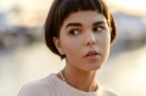 A person with short, dark hair and a straight fringe looks to the side. They wear a light-colored top and a beaded necklace. The background is softly blurred with warm, natural lighting.