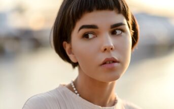 A person with short, dark hair and a straight fringe looks to the side. They wear a light-colored top and a beaded necklace. The background is softly blurred with warm, natural lighting.