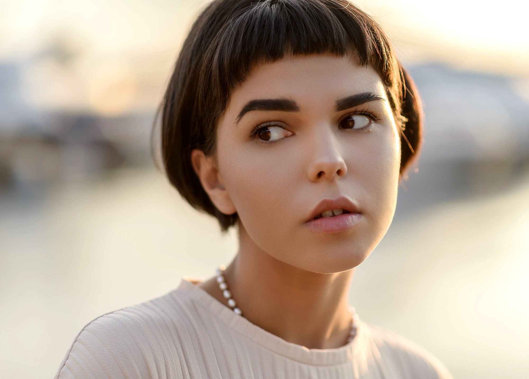 A person with short, dark hair and a straight fringe looks to the side. They wear a light-colored top and a beaded necklace. The background is softly blurred with warm, natural lighting.