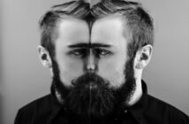 A bearded man in a black shirt stands in front of a mirror, creating a symmetrical reflection that blends his real image with the mirror. The black-and-white photograph emphasizes contrast and symmetry.