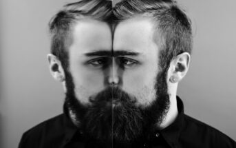 A bearded man in a black shirt stands in front of a mirror, creating a symmetrical reflection that blends his real image with the mirror. The black-and-white photograph emphasizes contrast and symmetry.