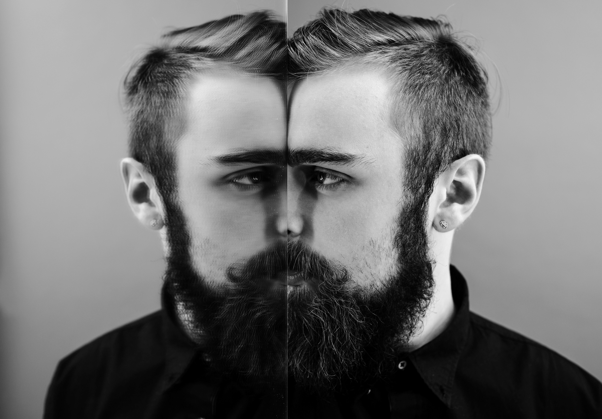 A bearded man in a black shirt stands in front of a mirror, creating a symmetrical reflection that blends his real image with the mirror. The black-and-white photograph emphasizes contrast and symmetry.