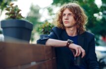 A person with long, curly hair sits on a wooden bench outdoors. They wear a casual blazer and hold a drink. Potted plants are on the bench, and the background features blurred greenery and soft lighting.