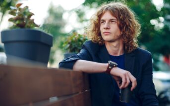 A person with long, curly hair sits on a wooden bench outdoors. They wear a casual blazer and hold a drink. Potted plants are on the bench, and the background features blurred greenery and soft lighting.