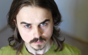 A person with long brown hair and a mustache is looking directly at the camera. They are wearing a green shirt and have a neutral expression. The background is softly blurred.