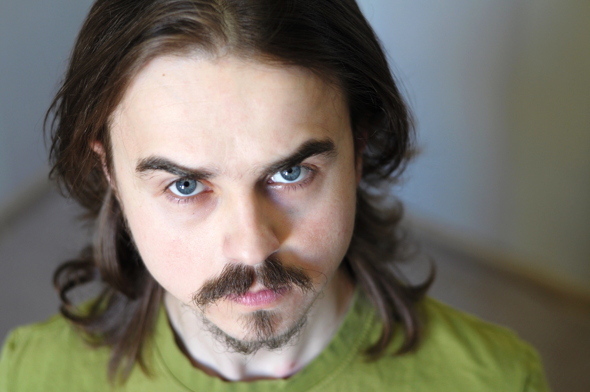 A person with long brown hair and a mustache is looking directly at the camera. They are wearing a green shirt and have a neutral expression. The background is softly blurred.