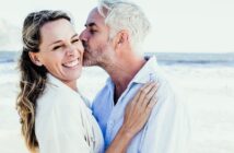 A man with gray hair kisses a smiling woman on the cheek at the beach. They both wear light-colored clothing, and the ocean is visible in the background, creating a joyful and serene atmosphere.