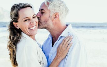 A man with gray hair kisses a smiling woman on the cheek at the beach. They both wear light-colored clothing, and the ocean is visible in the background, creating a joyful and serene atmosphere.