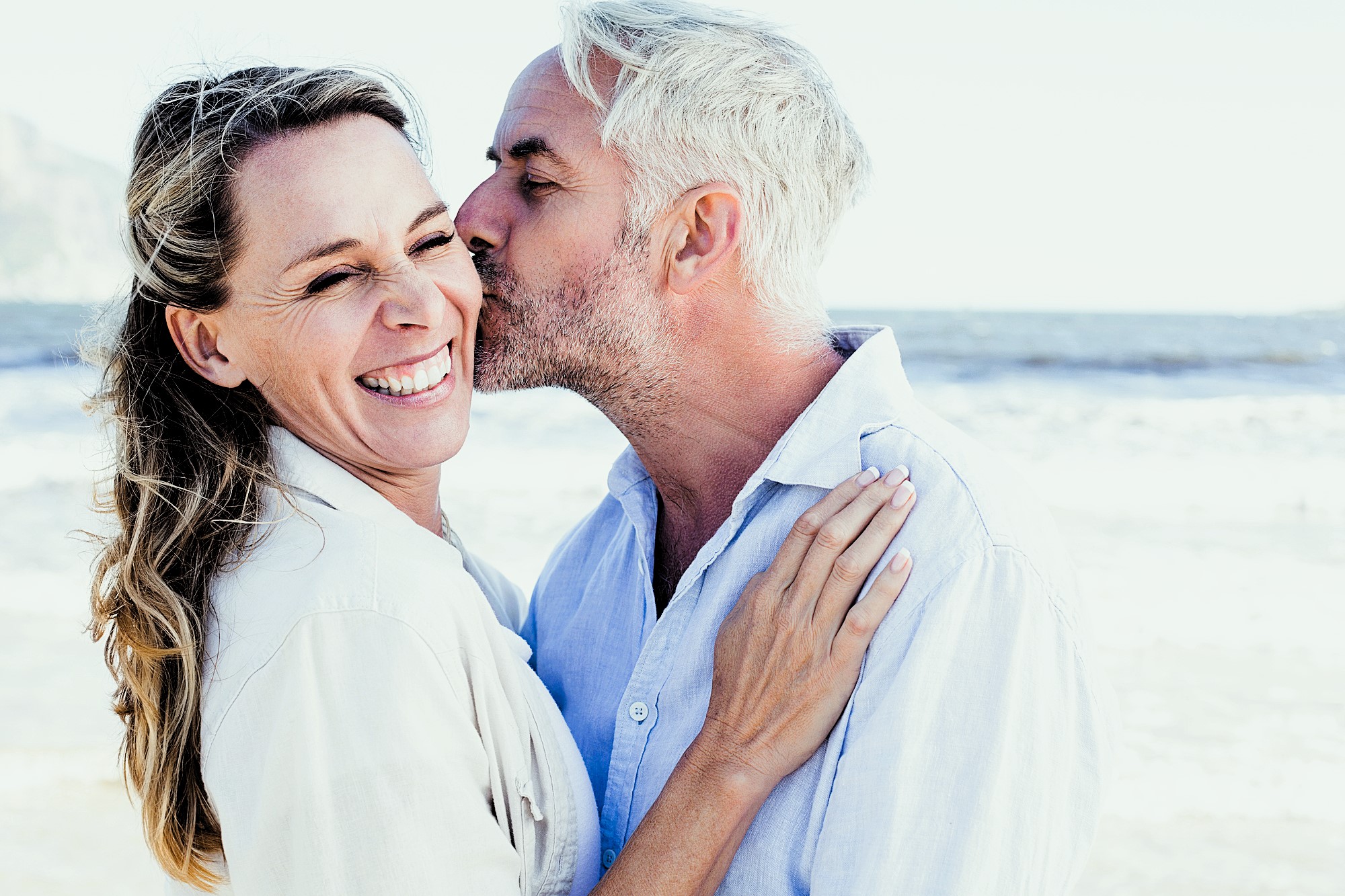 A man with gray hair kisses a smiling woman on the cheek at the beach. They both wear light-colored clothing, and the ocean is visible in the background, creating a joyful and serene atmosphere.