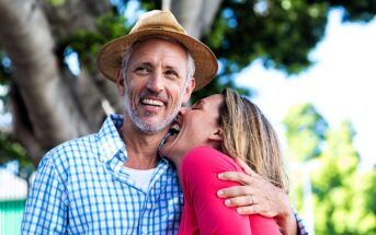 A cheerful couple is outdoors. The man, wearing a hat and a blue checkered shirt, smiles broadly while the woman in a pink top leans against him, laughing. They are surrounded by lush green trees.