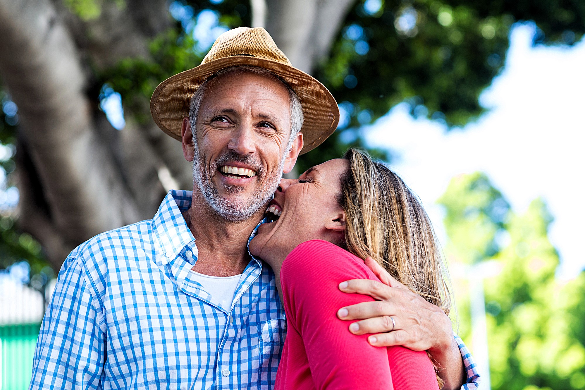 A cheerful couple is outdoors. The man, wearing a hat and a blue checkered shirt, smiles broadly while the woman in a pink top leans against him, laughing. They are surrounded by lush green trees.