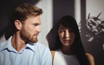 A man and a woman stand close together in dramatic lighting, with shadows of leaves cast on the wall behind them. The man has light hair and wears a blue shirt, while the woman has dark hair and wears a white top.