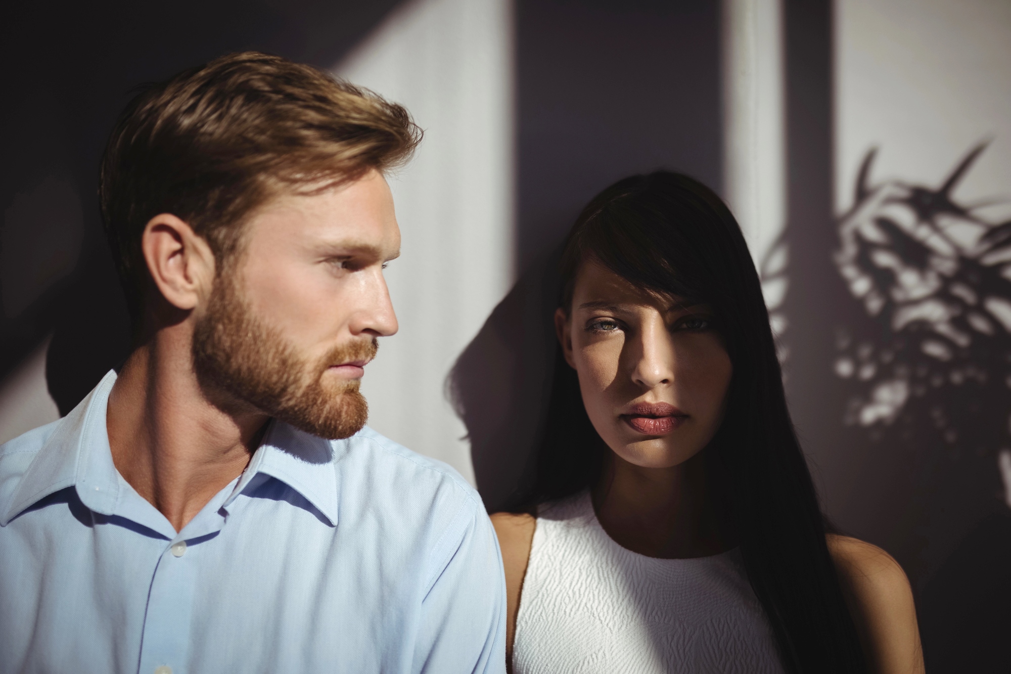 A man and a woman stand close together in dramatic lighting, with shadows of leaves cast on the wall behind them. The man has light hair and wears a blue shirt, while the woman has dark hair and wears a white top.