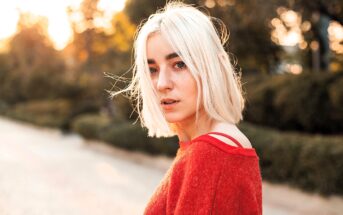 A young person with short, blonde hair stands outdoors. They are wearing a red sweater and looking at the camera. The background is softly blurred, with warm sunlight filtering through trees.